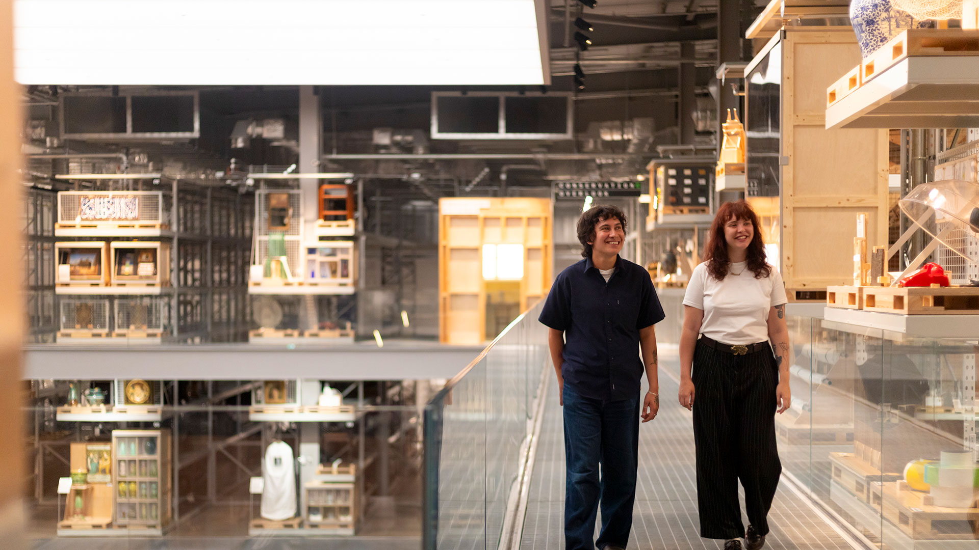 View of Weston Collections Hall at V&A East Storehouse. Image courtesy of Kemka Ajoku/V&A East Storehouse. A man and a woman walking round an elevated floor of the storehouse with artefacts on display around them