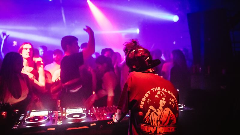 Corsica Studios was one of London's first warehouse venues. Credit: Sienna Lorraine Gray. Image courtesy of Corsica Studios. People dancing in front of female DJ spinning tunes at Corsica Studios nightclub under purple and red lights.
