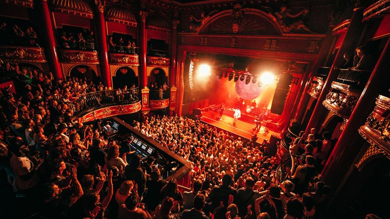 Discover great live music acts at KOKO. Credit: Phoebe Fox. Image courtesy of KOKO. A crowd of people facing a brightly lit stage at the KOKO club in London.