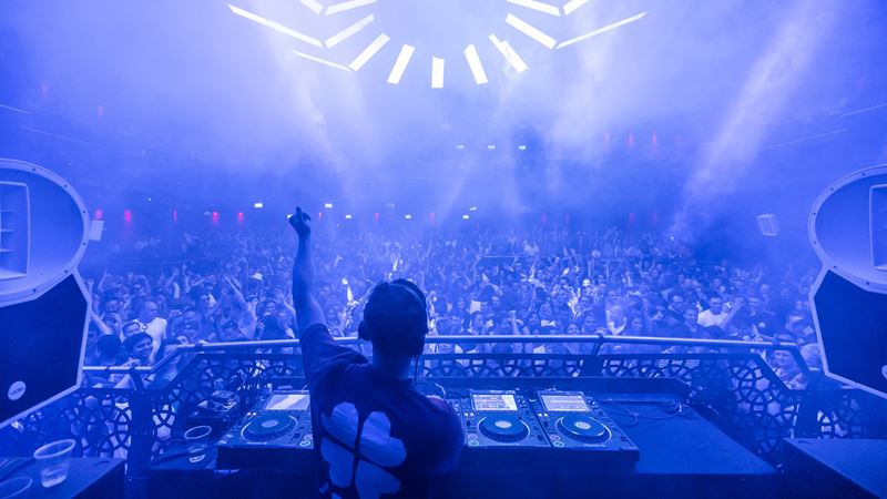 DJ spinning tunes with one arm up in the air in front of a crowd dancing in a nightclub lit up in blue.