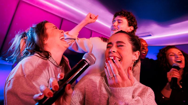 Make memories with music at Lucky Voice karaoke. Credit: Ant Tran. Image courtesy of Lucky Voice Group. A photo of five friends laughing and singing into a microphone at Lucky Voice karaoke with pink neon lights illuminating the room
