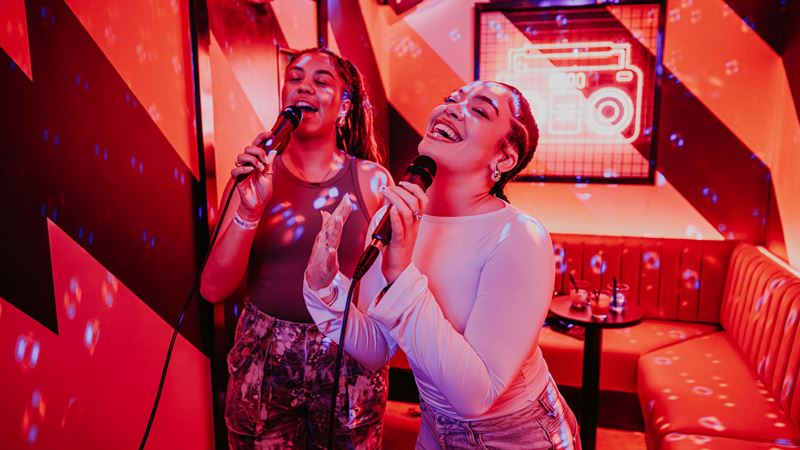 Singalong to your favourite tunes at PLAYBOX in Boxpark Croydon. Credit: Luke Dyson. Image courtesy of PLAYBOX. A photo of two girls smiling and singing into microphones inside a karaoke booth at Playbox with red lighting
