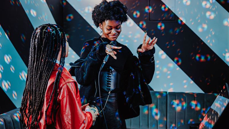 A photo of two girls dancing along to music inside a karaoke booth as one girl sings into a microphone with bubbles in the air