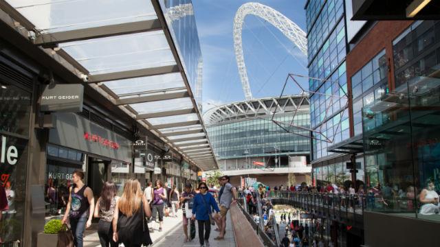 People are shopping and relaxing in a bright and modern shopping centre, partially open to the outdoors on a sunny day.