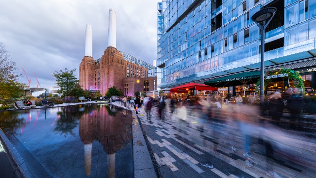 The outside of Battersea Power Station reflected in some water with restaurants and blurred people in the foreground.