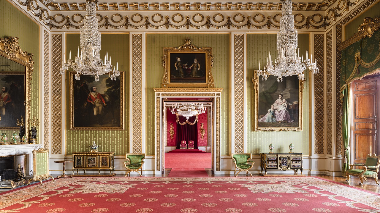 Landscape photo of the Green Drawing Room in Buckingham Palace; royal gold and red carpet, green panelled walls with large portraits hanging, high ceilings decorated in gold, green and gold furniture in the room