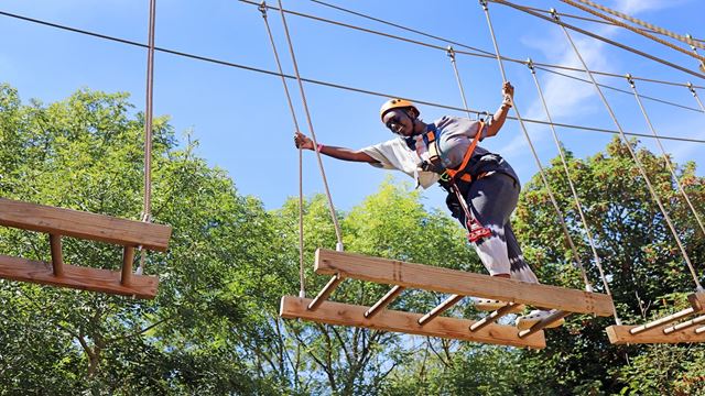 a young woman climbing on a high ropes course, smiling as she reaches for the next platform to climb onto