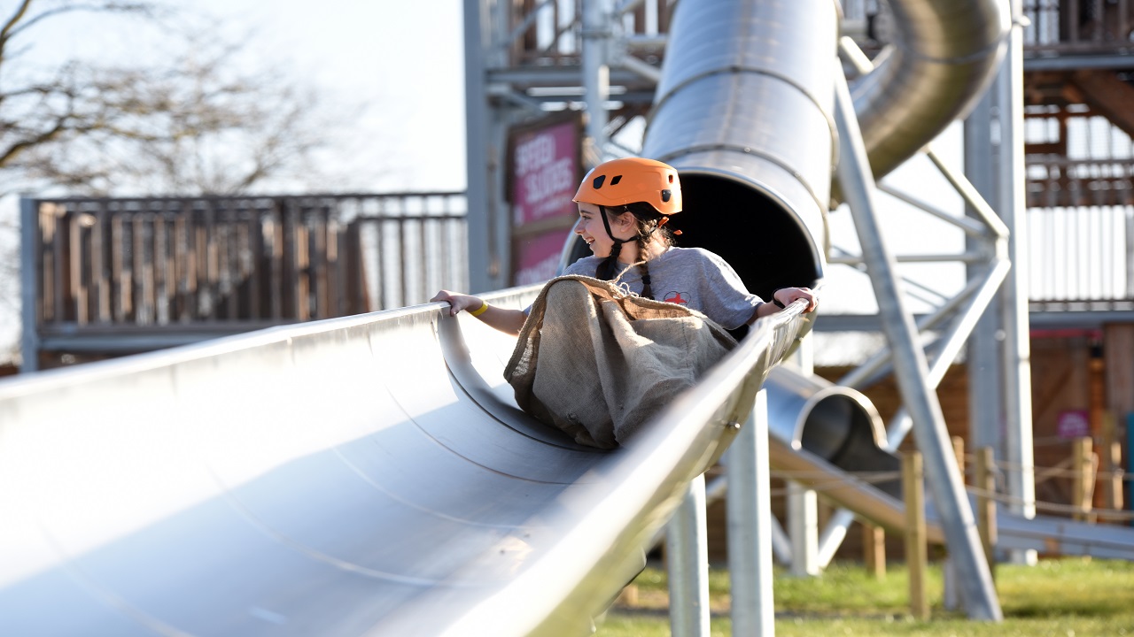 Zoom down a 24-metre high speed slide at Gripped London. Image courtesy of Golden Tours. a young girl wearing an orange helmet and sat in a sack, sliding down a tall, steel covered slide