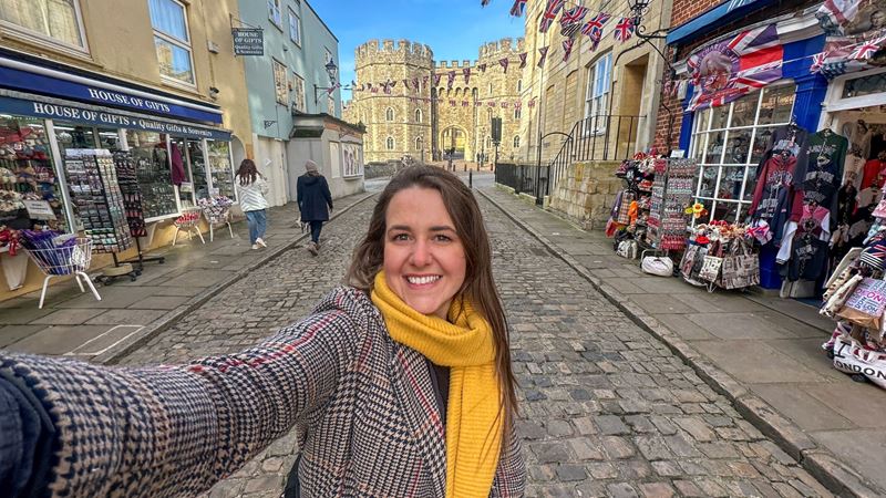 A woman in a brown coat and yellow scarf takes a selfie in front of windsor castle.