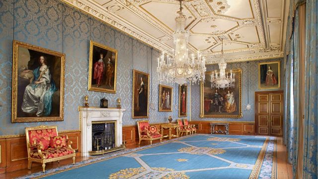 A blue room with blue carpets and red sofas with paintings along the wall at the queens gallery at windsor castle.