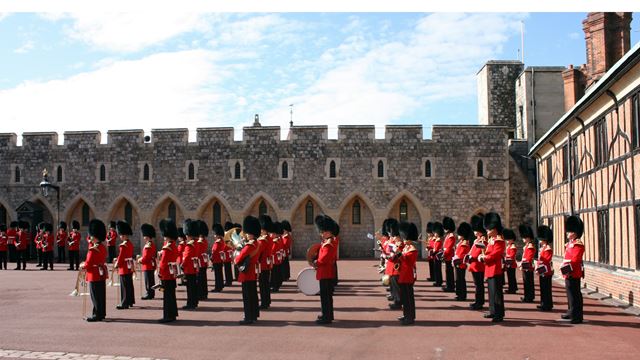 Guards dressed in red with black hats in formation at Windsor Castle on a sunny day.