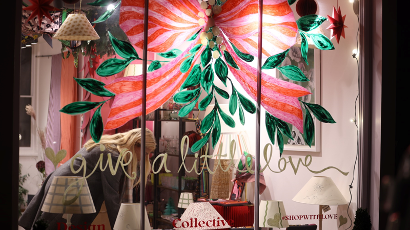 Venture to Connaught Village for a spot of Christmas shopping. Credit: Dave Parry, PA. Image courtesy of Connaught Village. A view of a shop window from outside with a giant pink Christmas bow mural with a woman looking through items inside