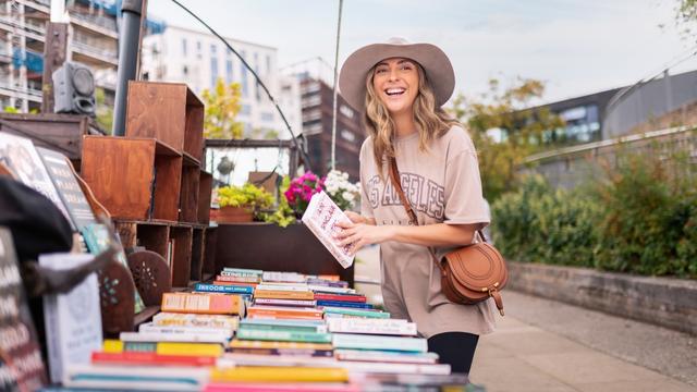 Seek out fantastic souvenirs at local London markets. © visitlondon/Antoine Buchet A girl with a tshirt and brown bag wearing a hat stands holding a book on a barge market stall in london.