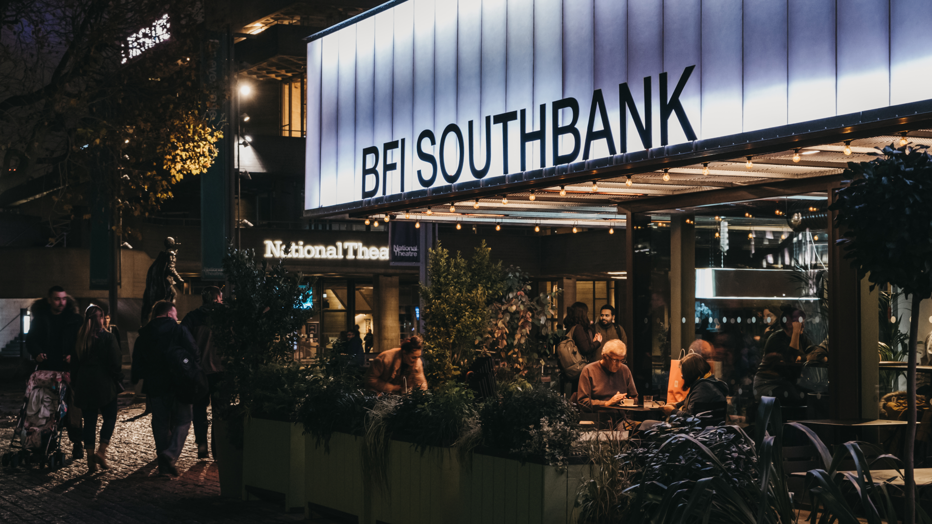 Exterior of BFI Southbank pictured at night. An image of the front of BFI Southbank at night with groups of people sat outside at tables and walking in front of the building with lights illuminating the venue
