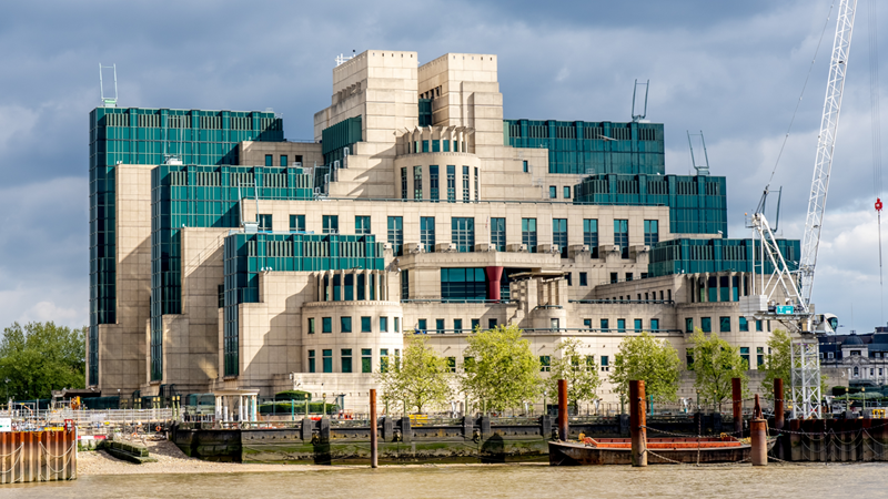 Catch a glimpse of Thames House, the home of the British Secret Intelligence Service and a feature in many Bond films. Credit: Shutterstock. Image courtesy of Shutterstock. A photo of Thames House on a sunny day with the river Thames in the foreground, featuring beige brick and blue coloured glass features