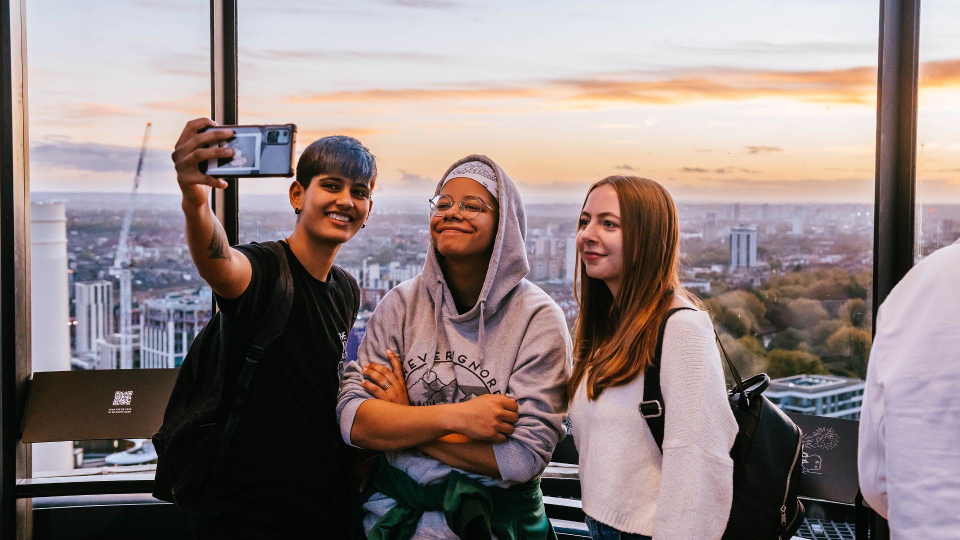 Snap a selfie with epic views of London at Lift 109 at Battersea Power Station. Image courtesy of The Cast. Photo Credit: Joshua Atkins Three people take a selfie in lift 109 at battersea power station with the london skyline at sunset as the backdrop.