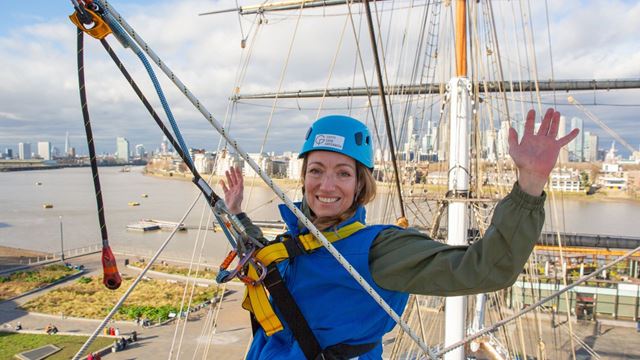 a woman wearing a jumpsuit and hard hat, smiling and waving at the top of the Cutty Sark, with a view of the London skyline behind