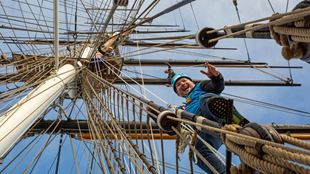 a photo taken from the deck of the Cutty Sark, looking towards the sky with people harnessed and climbing the Cutty Sark, waving and smiling