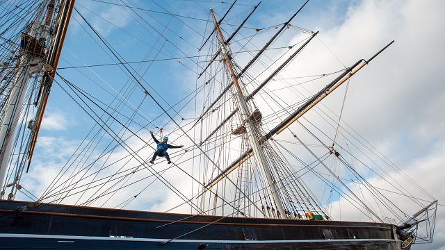 a low angle photo taken from a distance of a person climbing the Cutty Sark, wearing a jumpsuit and harness