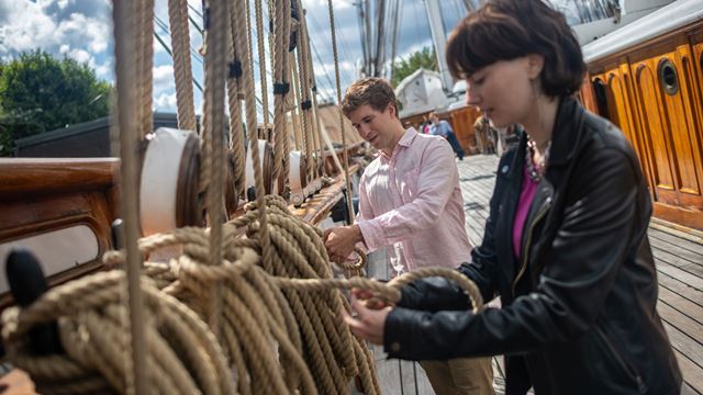 A man and a woman are on the upper deck of the Cutty Sark trying their hand at marine rope knots. 