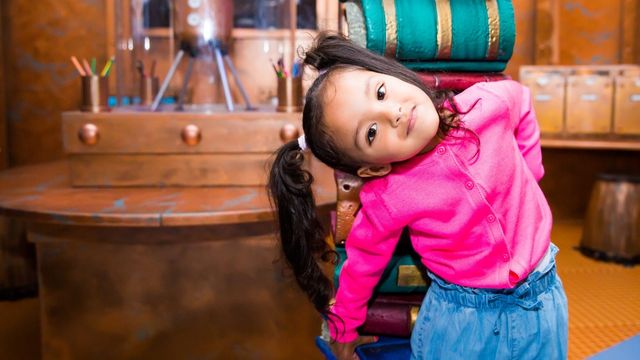 A toddler in a bright pink jumper and blue trousers stands against a pillar made of books.