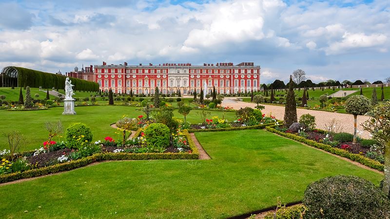 Walk through the magnificent gardens of Hampton Court Palace. Credit: Shutterstock. Image courtesy of Shutterstock. A long walkway leads to the brick Tudor Palace of Hampton Court, borered by flower beds and French-inspired landscaping.
