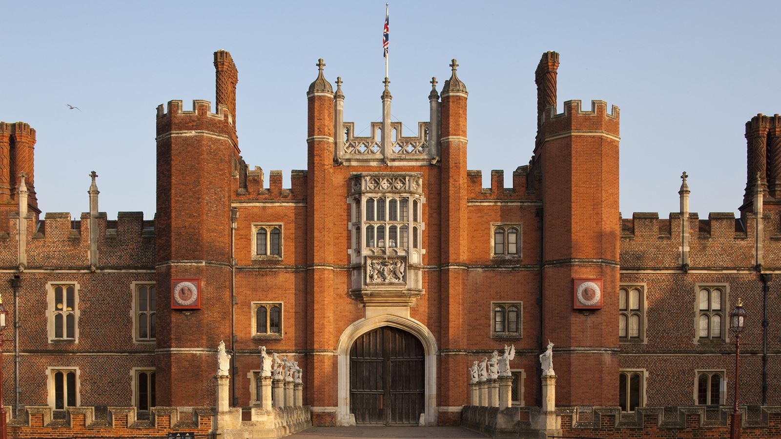 The ornate red brick and turreted facade of the West Front of Hampton Court Palace on a clear day.