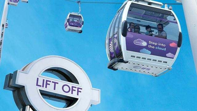 The purple IFS Cloud cable cars fly over the Thames in London. Image courtesy of TfL. The purple coloured IFS Cloud Cable Cars fly over a Transport for London sign with the words Lift Off on it.