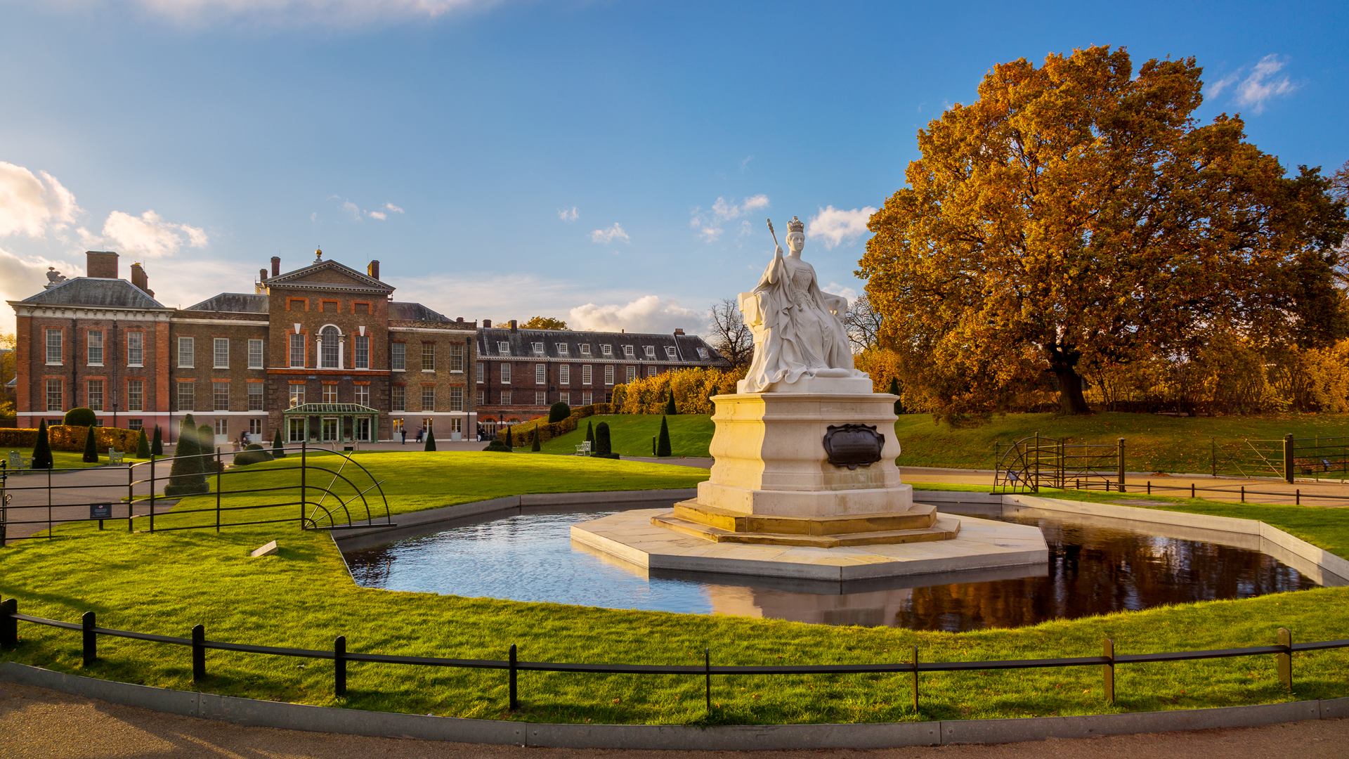 Discover Kensington Gardens and its myriad things to see and do. Credit: Jon Reid. Image courtesy of visitlondon.com. Queen Victoria statue in Kensington Gardens, with tree with autumn leaves, and Kensington Palace in the background