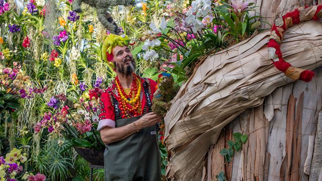 A man wearing an assortment of colourful necklaces and a green stuffed lizard on his head stands next to floral installations, part of the Orchid Festival at Kew Gardens.