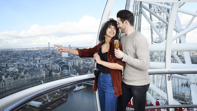 A male and female couple hold a glass of champagne as they stand in a London Eye capsule, with London's skyline in the background.