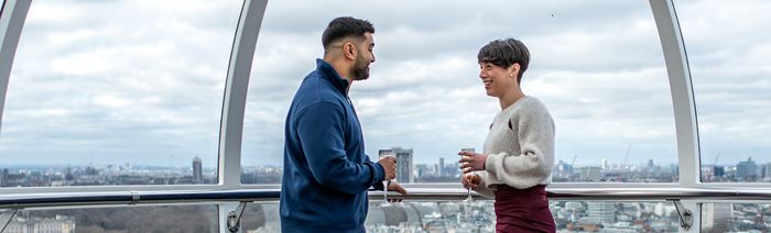 A couple hold glasses of champagne aboard the London Eye, with views of London's skyline in the background.