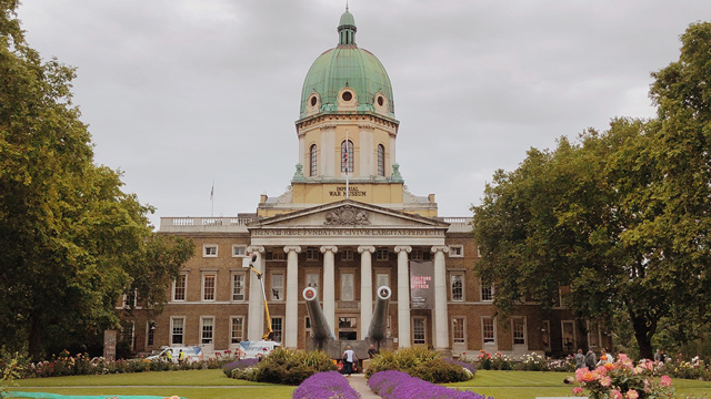 A tall white building with a green dome and five pillars. Surrounded by trees.