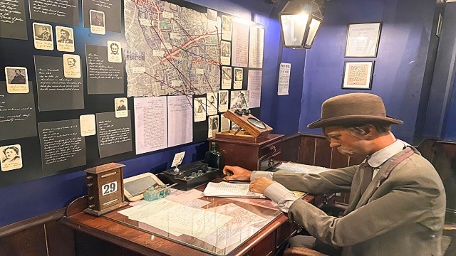 A male mannequin wearing Victorian clothes, sat at a wooden desk with profile sketches and a map on the wall in front of them.