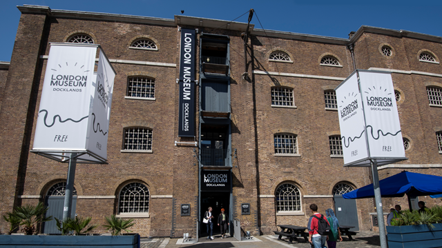 The front exterior of the London Museum Docklands with two large signs outside displaying the museum's logo