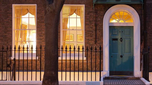 The exterior of the Charles Dickens Museum with gated railings, two large windows showing the rooms lit up inside and a green front door