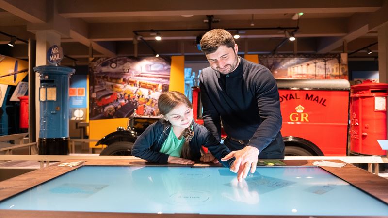 Explore interactive exhibitions and learn about the fascinating history of mail. Image courtesy of The Postal Museum. A man and a young child looking at an interactive display table with mail memorabilia in the background