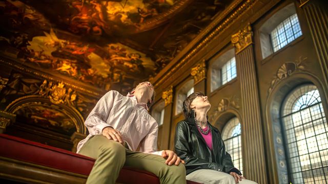 Two people look at at the ceiling in the Painted Hall, which is decorated in colourful frescoes, within the Old Royal Naval College.