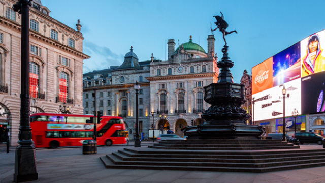 Piccadilly Circus at dusk, with the statue of Eros in the foreground, and a red London bus and the iconic wall of LED lights in the background. 