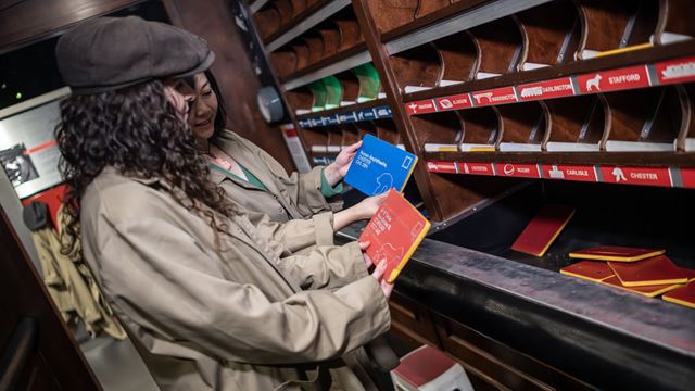 Two women, dressed with trench coats and grey flat caps, are sorting out mail at the Postal Museum. 