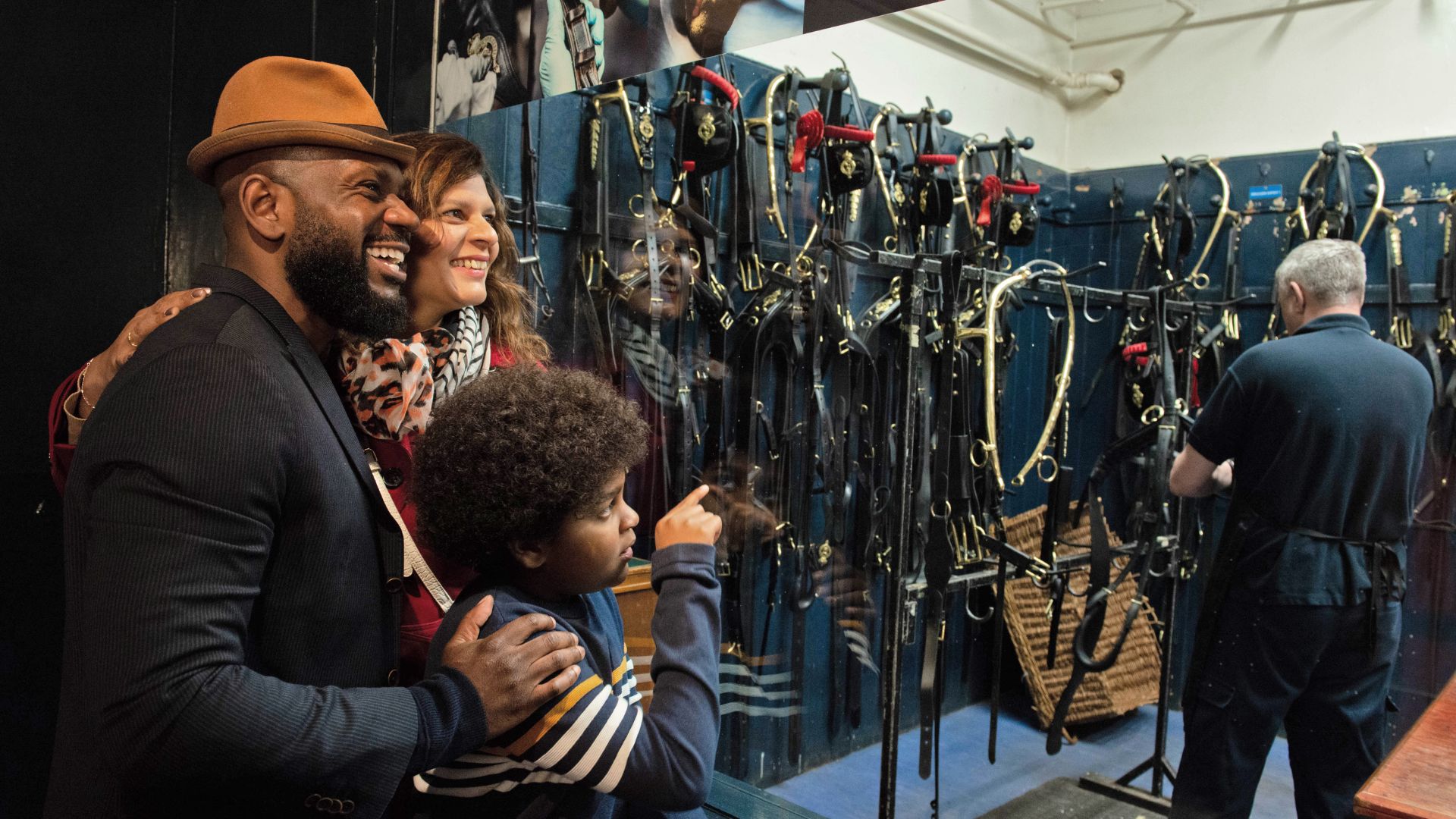 Inside the Harness Room at the Royal Mews, where visitors can see the elaborate leather harnesses and fittings used to prepare horses for state occasions. © Royal Collection Enterprises Limited 2025 | Royal Collection Trust Family viewing the Harness Room display of royal harnesses and tack, with a staff member working in the background at the Royal Mews.