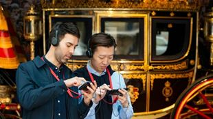 Two people stand next to a gold and black carriage listening to an audio guide.