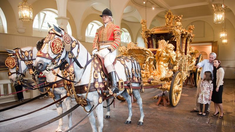 Family viewing the Gold State Coach at the Royal Mews, with model horses and riders in ceremonial harnesses on display.