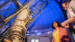 Two people stand under the large white telescope under blue light at the royal observatory greenwich.