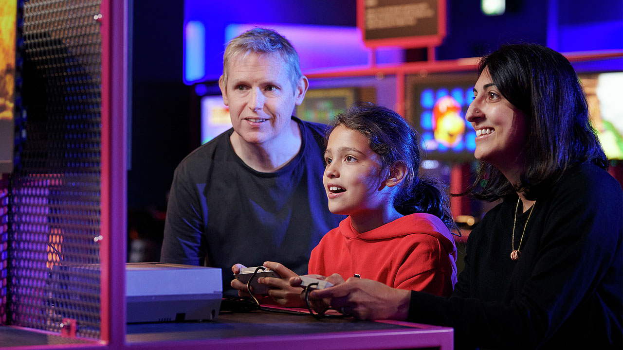 Power Up at the Science Museum. Image courtesy of the Science Museum Group. Family crowded round games station with child holding games console.