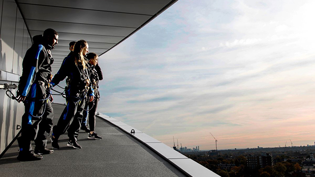 The Dare Skywalk at Tottenham Hotspur Stadium. Image courtesy of Golden Tours. A small group of people, attached to a railing, peer over the edge of Tottenham Hotspur Stadium at sunset, with London's skyline in the background.
