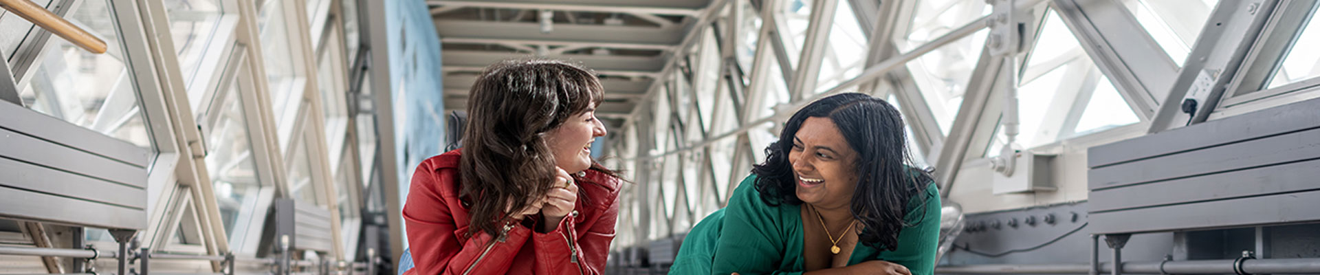 Two women are laughing together while looking down the glass walkway at Tower Bridge. 
