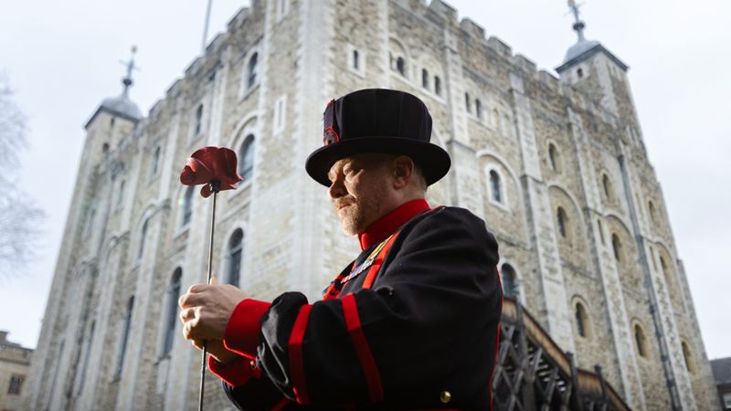 A yeoman warder holds a ceramic poppy in front of the tower of london.