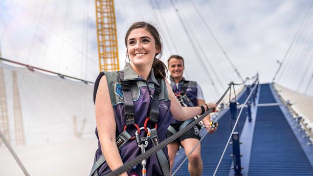 A woman and man walk down the side of The O2, hanging onto a rope and wearing harnesses, during the Up at The O2 experience.