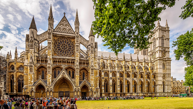 A wide-angle shot of Westminster Abbey on a sunny day, taken from across Parliament Square, with a crowd in the foreground.